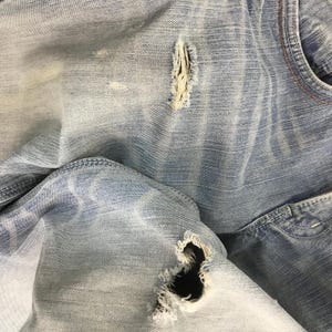 May include: Close-up of light blue denim jeans with distressed details. The jeans have multiple tears and frayed edges, showcasing a worn and vintage aesthetic. The fabric displays subtle fading and texture variations.