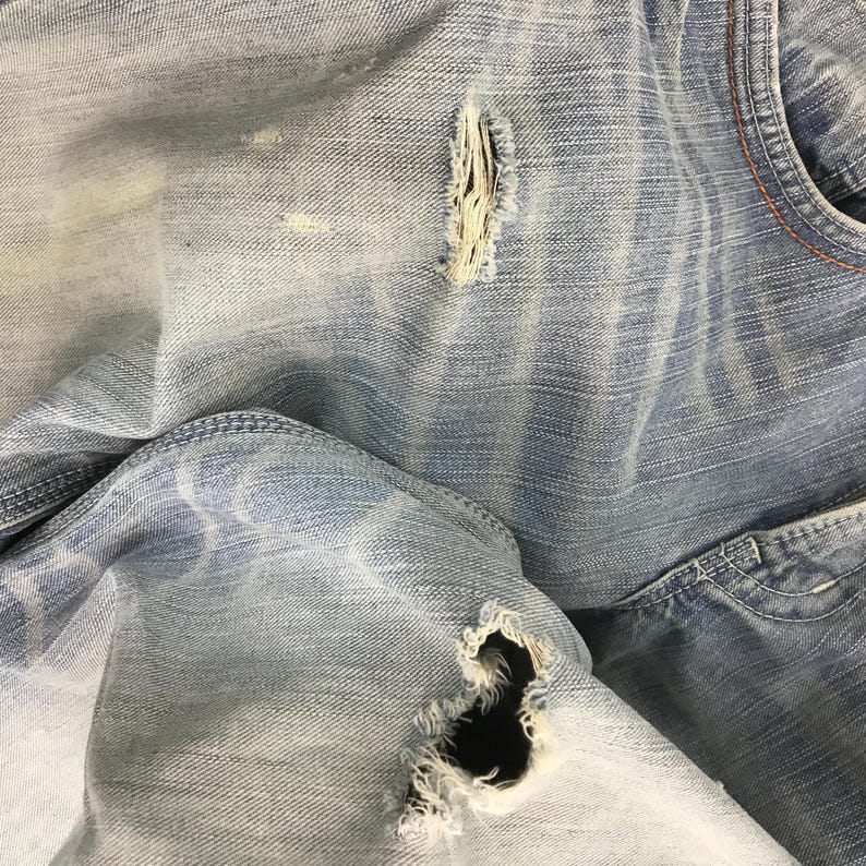 May include: Close-up of light blue denim jeans with distressed details. The jeans have multiple tears and frayed edges, showcasing a worn and vintage aesthetic. The fabric displays subtle fading and texture variations.