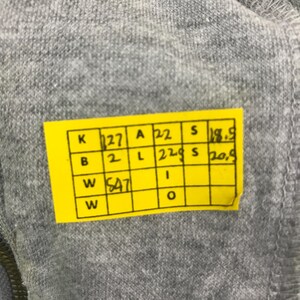 May include: Close-up of gray fabric with a yellow tag attached. The tag has black text and numbers in a grid format. The fabric appears to be a soft material.