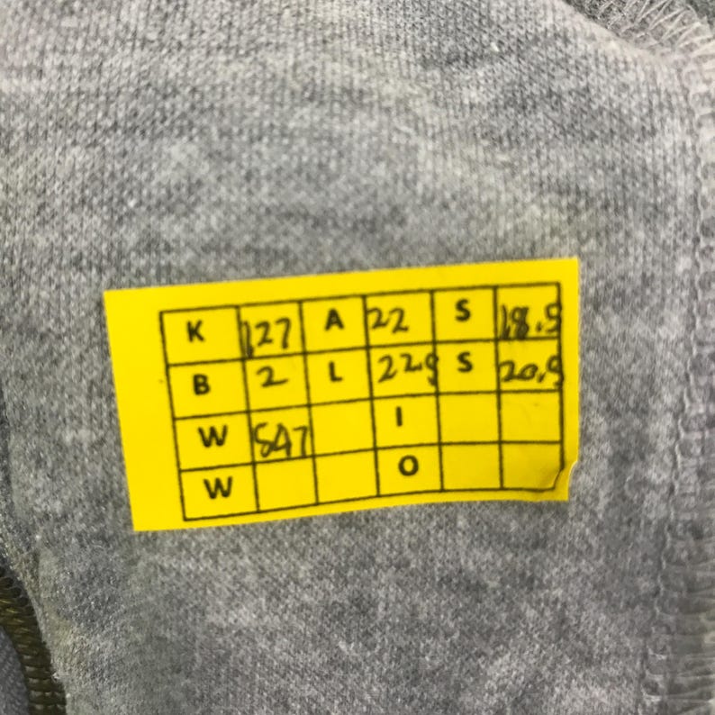 May include: Close-up of gray fabric with a yellow tag attached. The tag has black text and numbers in a grid format. The fabric appears to be a soft material.
