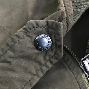 May include: An olive green jacket featuring a dark metal snap button embossed with the text AVIREX U.S.A. and a registered trademark symbol. The garment includes a ribbed collar, visible stitching along the lapel, and a metal zipper pull.