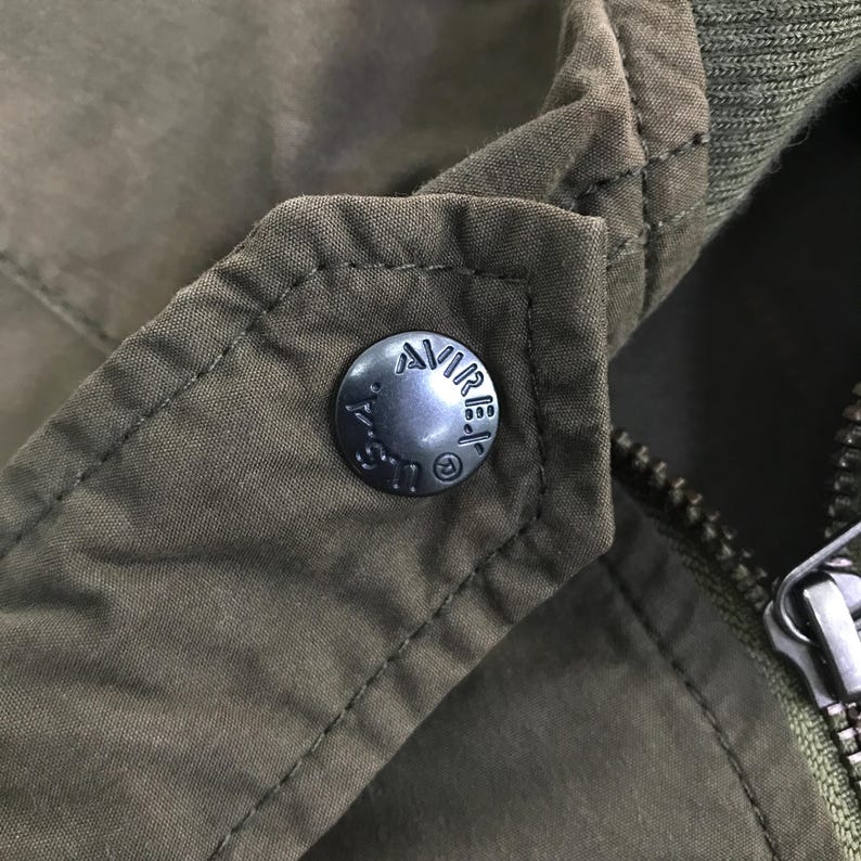 May include: An olive green jacket featuring a dark metal snap button embossed with the text AVIREX U.S.A. and a registered trademark symbol. The garment includes a ribbed collar, visible stitching along the lapel, and a metal zipper pull.