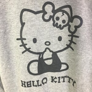 May include: A gray sweatshirt featuring a black graphic of Hello Kitty with a skull and crossbones bow. The words "HELLO KITTY" are printed in a block font at the bottom of the design.