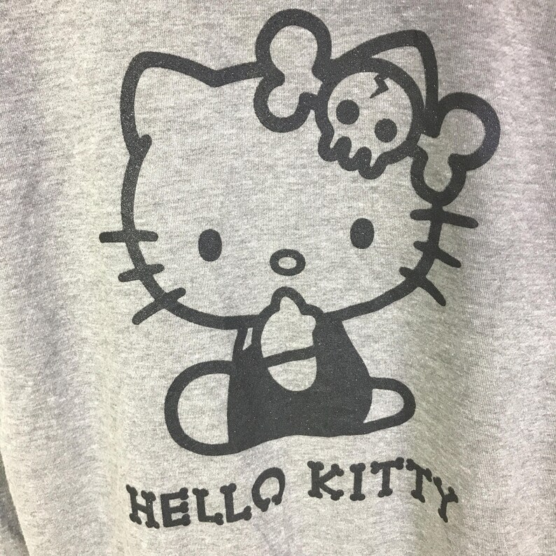 May include: A gray sweatshirt featuring a black graphic of Hello Kitty with a skull and crossbones bow. The words "HELLO KITTY" are printed in a block font at the bottom of the design.