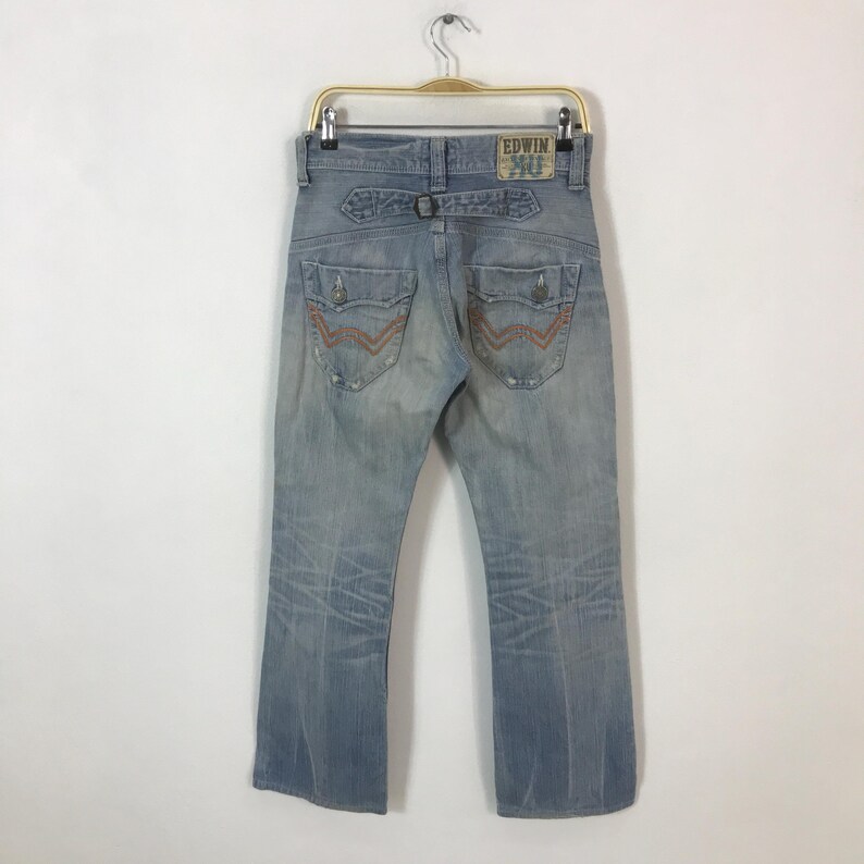 May include: Light blue denim jeans with distressed details, featuring a back belt loop and pockets with orange stitching. The jeans have a faded wash and the "EDWIN 40" logo patch.
