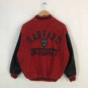 以下が含まれることがあります： 黒い革の袖が付いた赤いハーバード大学のレターマンジャケット。ジャケットの背面にはハーバード大学のロゴとテキストがあります。木製のハンガーに吊るされています。