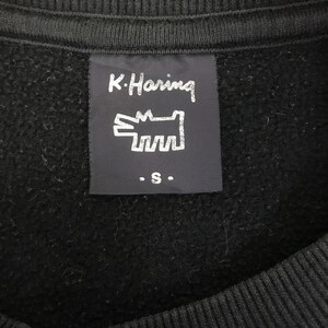 K&ouml;nnte beinhalten: Nahaufnahme eines schwarzen Sweatshirts mit einem Etikett mit dem Text "K. Haring" und einer stilisierten Hunde-Grafik. Das Etikett enth&auml;lt auch den Buchstaben "S". Das Sweatshirt besteht aus einem weichen, strukturierten Material.