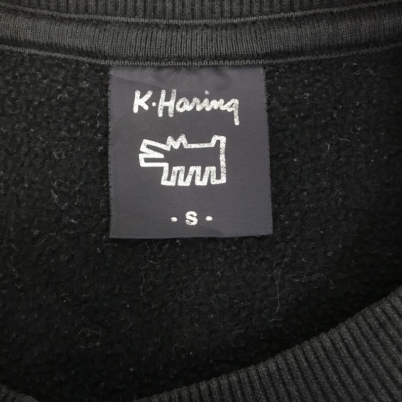 K&ouml;nnte beinhalten: Nahaufnahme eines schwarzen Sweatshirts mit einem Etikett mit dem Text "K. Haring" und einer stilisierten Hunde-Grafik. Das Etikett enth&auml;lt auch den Buchstaben "S". Das Sweatshirt besteht aus einem weichen, strukturierten Material.