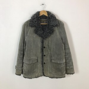 Puede incluir: Chaqueta de pana gris con cuello y ribete de piel sintética. La chaqueta tiene cierre de botones, dos bolsillos delanteros y puños abotonados. Colgada de una percha de madera.