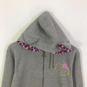 May include: A gray zip-up hoodie with a hood featuring cat ears and a pink and black leopard print lining. The front of the hoodie has a gold outline of a cat face with a pink bow.
