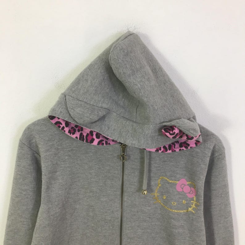 May include: A gray zip-up hoodie with a hood featuring cat ears and a pink and black leopard print lining. The front of the hoodie has a gold outline of a cat face with a pink bow.