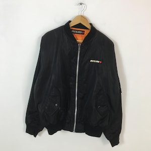 May include: Black bomber jacket with the word "nismo" in white and red on the chest and collar. The jacket has a zipper closure, two front pockets, and ribbed cuffs and hem. The interior lining is orange.