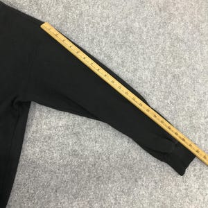 K&ouml;nnte beinhalten: Schwarzes Langarm-Sweatshirt mit einem Holzlineal auf dem &Auml;rmel. Das Lineal ist in Zoll markiert und zeigt ein Ma&szlig; von etwa 58 cm. Das Sweatshirt liegt auf einer grauen Oberfl&auml;che.