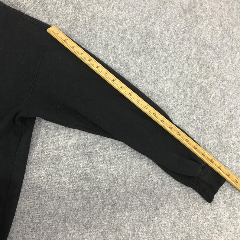 K&ouml;nnte beinhalten: Schwarzes Langarm-Sweatshirt mit einem Holzlineal auf dem &Auml;rmel. Das Lineal ist in Zoll markiert und zeigt ein Ma&szlig; von etwa 58 cm. Das Sweatshirt liegt auf einer grauen Oberfl&auml;che.