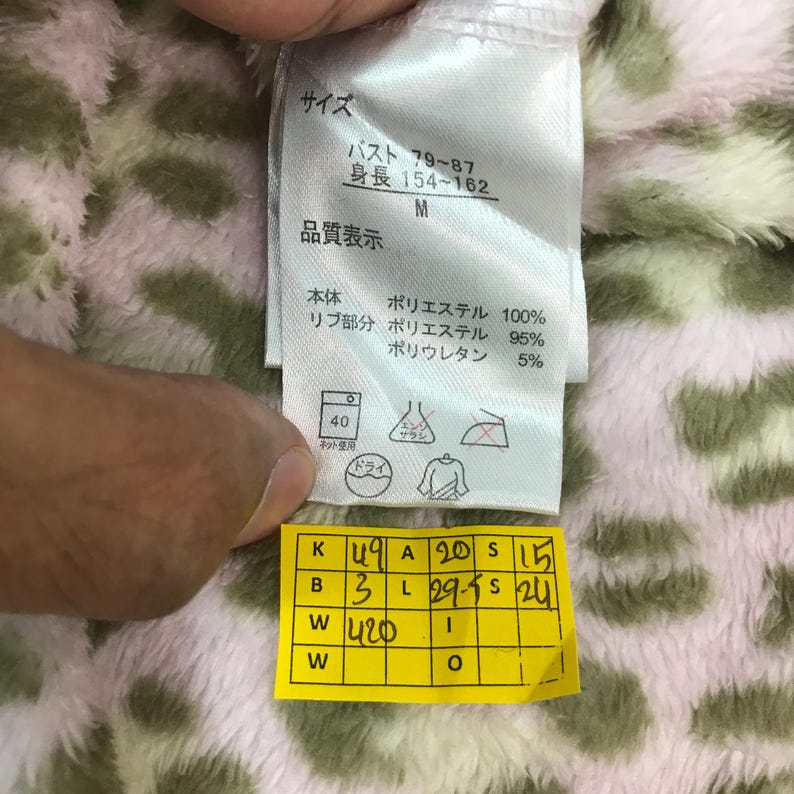 Puede incluir: Primer plano de una etiqueta de ropa con texto japon&eacute;s y s&iacute;mbolos de cuidado. La etiqueta incluye medidas de busto y altura, junto con detalles de la composici&oacute;n de la tela. Una etiqueta amarilla con n&uacute;meros y letras est&aacute; adjunta.