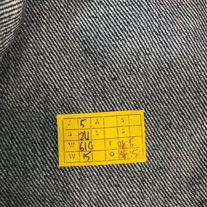May include: Close-up of blue denim fabric with a yellow tag. The tag has several numbers and letters printed on it, including "5", "21", "610", and "15". The denim has a diagonal weave pattern.