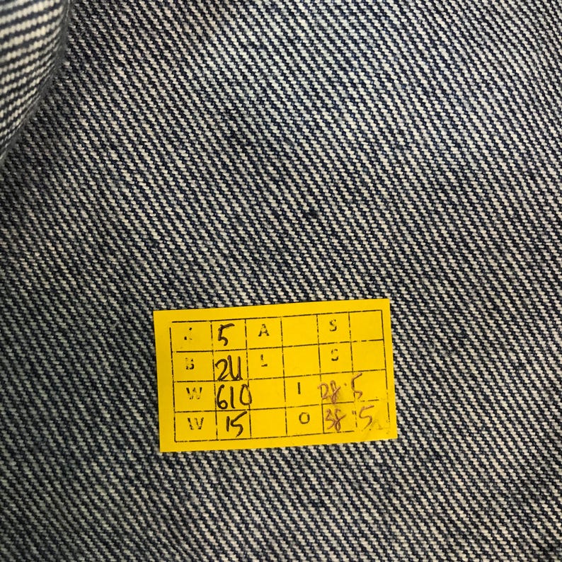 May include: Close-up of blue denim fabric with a yellow tag. The tag has several numbers and letters printed on it, including "5", "21", "610", and "15". The denim has a diagonal weave pattern.