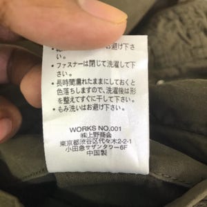 May include: A white rectangular garment care label with black Japanese text printed on it. The label includes instructions to close zippers before washing, avoid soaking, and reshape before drying. It lists WORKS NO.001 and company details.