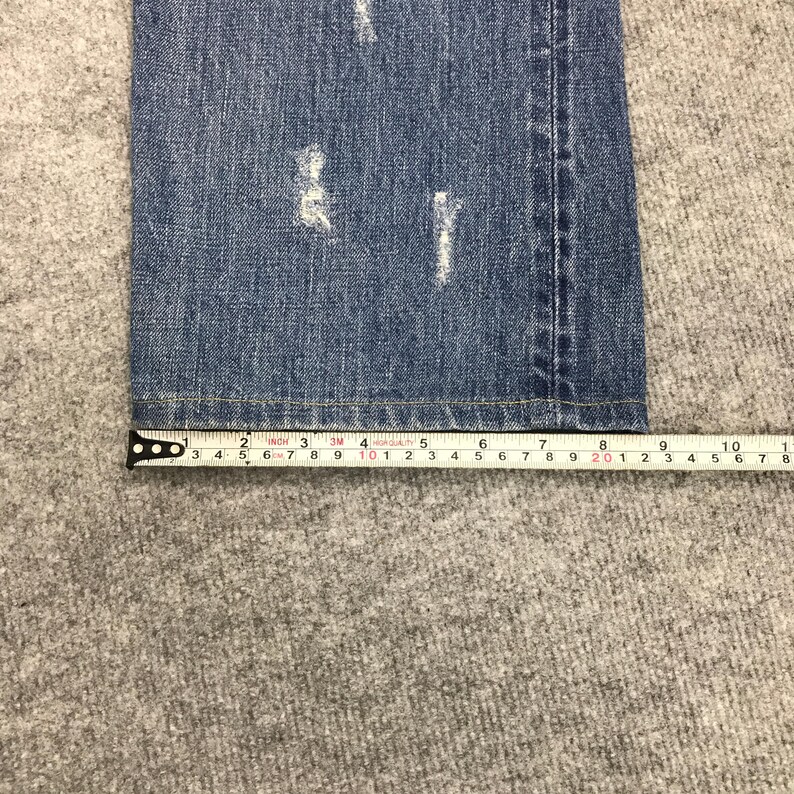 May include: Close-up of blue denim jeans with distressed details. A measuring tape shows the jeans are approximately 10 inches wide. The jeans are on a gray surface.