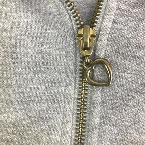 May include: Close-up of a gray hoodie zipper with a gold-colored metal zipper pull. The zipper pull is in the shape of a heart. The zipper is closed.