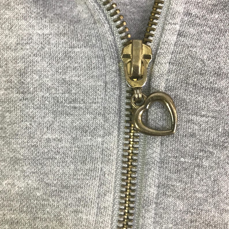 May include: Close-up of a gray hoodie zipper with a gold-colored metal zipper pull. The zipper pull is in the shape of a heart. The zipper is closed.