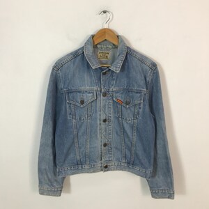 May include: A light blue denim jacket with a classic collar, button-down front, and two chest pockets. The jacket has a faded wash and a small red logo patch. It is hanging on a wooden hanger.