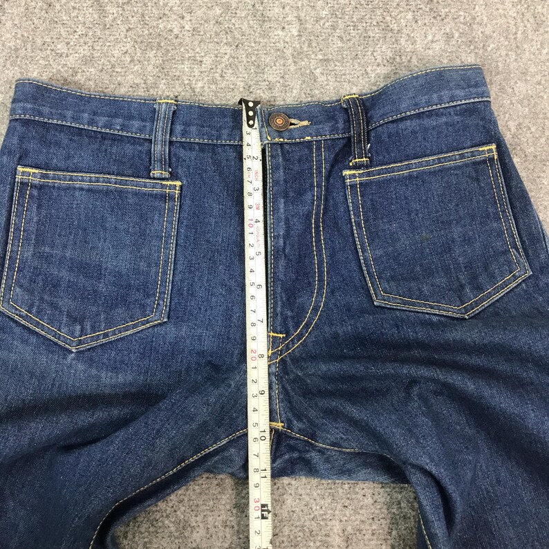 May include: Close-up of blue denim jeans with yellow stitching. A measuring tape shows the waist is approximately 10 inches wide. The jeans have two front pockets and two back pockets.