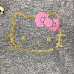 May include: A gray fabric with a gold glitter outline of a cartoon cat face. The cat has a pink bow and gold glitter eyes and nose. The design is centered on the fabric.