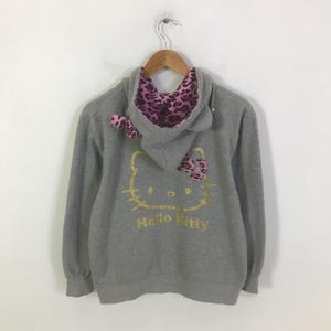 May include: A gray hooded sweatshirt featuring a gold Hello Kitty graphic on the back. The hood lining and bow are pink with a leopard print. The sweatshirt is hanging on a wooden hanger.