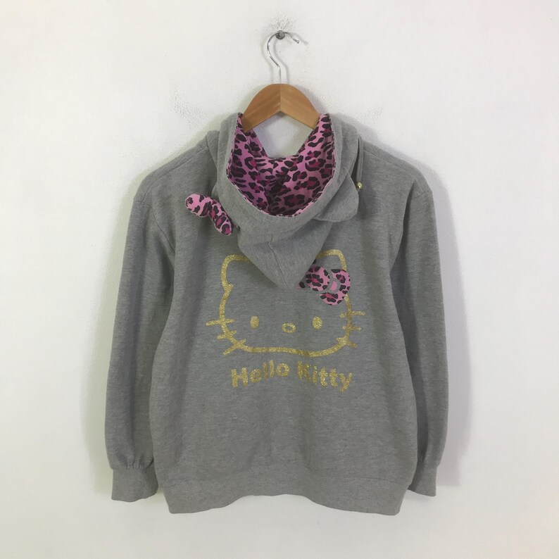May include: A gray hooded sweatshirt featuring a gold Hello Kitty graphic on the back. The hood lining and bow are pink with a leopard print. The sweatshirt is hanging on a wooden hanger.