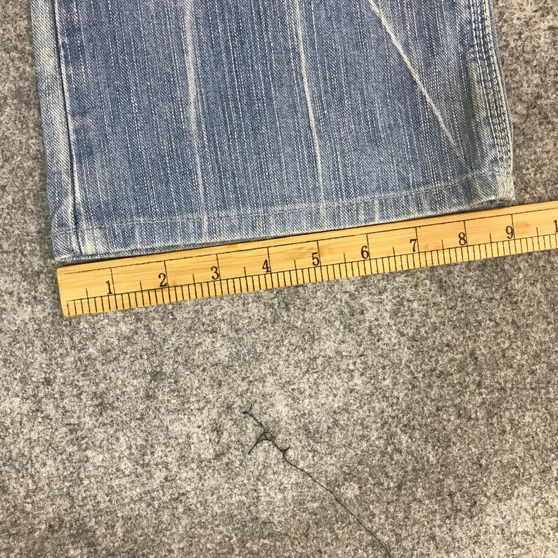 May include: Close-up of light blue denim jeans with a wooden ruler placed along the hem. The ruler shows measurements in inches. The jeans have visible seams and a faded appearance, suggesting wear.