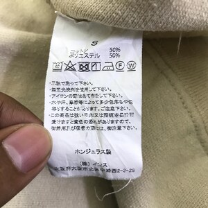 May include: A close-up of a clothing label with Japanese text and care symbols. The label states the garment is 50% cotton and 50% polyester. Washing instructions are also included, along with the manufacturer's address in Osaka, Japan.