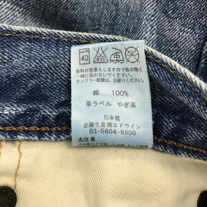 May include: Close-up of a blue denim jeans label with Japanese text and care symbols. The label indicates the jeans are 100% cotton and made in Japan. The label also includes washing instructions and a phone number.