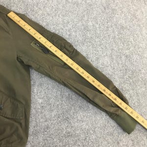 May include: An olive green bomber jacket sleeve featuring a zippered utility pocket on the upper arm and a ribbed cuff. A wooden ruler is placed along the sleeve, showing a length measurement of approximately 25 inches from shoulder to cuff.