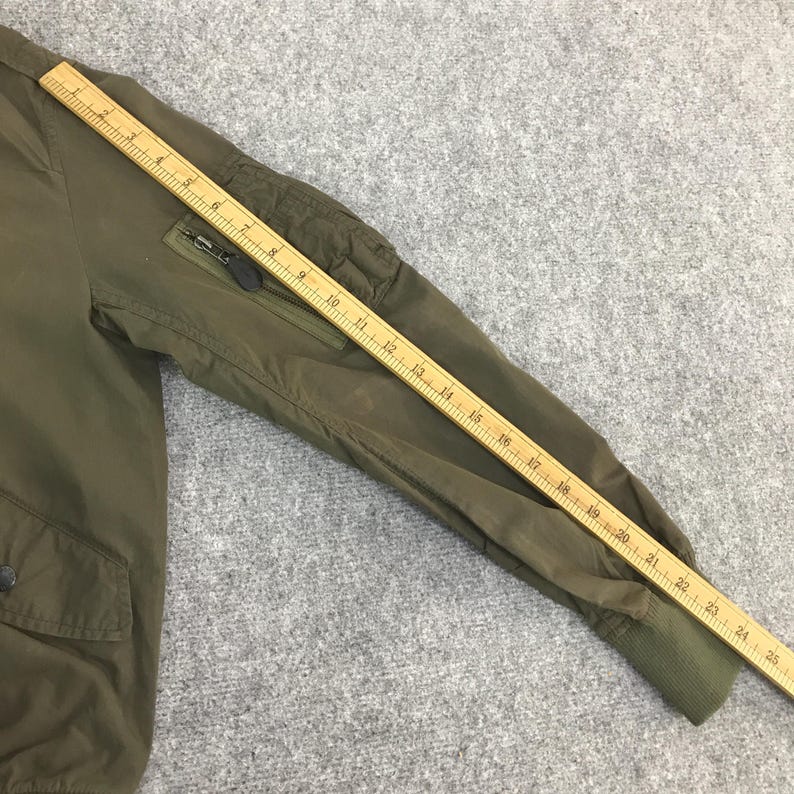 May include: An olive green bomber jacket sleeve featuring a zippered utility pocket on the upper arm and a ribbed cuff. A wooden ruler is placed along the sleeve, showing a length measurement of approximately 25 inches from shoulder to cuff.
