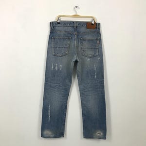 May include: Light blue denim jeans with distressed details, including faded areas and rips. The jeans have a classic five-pocket design, a leather patch, and are hanging on a hanger. The jeans are a vintage style.