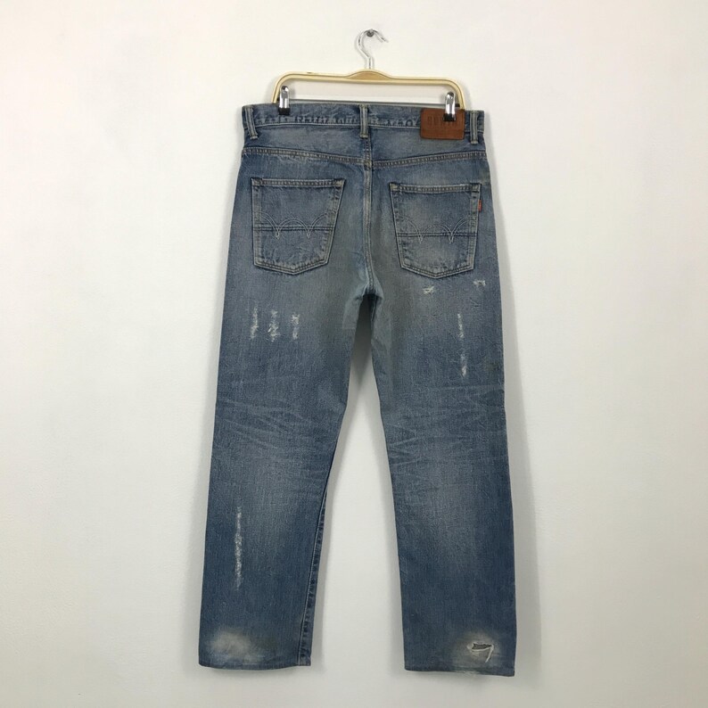 May include: Light blue denim jeans with distressed details, including faded areas and rips. The jeans have a classic five-pocket design, a leather patch, and are hanging on a hanger. The jeans are a vintage style.