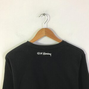 K&ouml;nnte beinhalten: Schwarzes Sweatshirt mit Rundhalsausschnitt, das an einem Holzb&uuml;gel h&auml;ngt. Auf der R&uuml;ckseite des Shirts steht der wei&szlig;e Text "&copy; K. Haring" in handschriftlicher Form. Das Shirt besteht aus einem dunklen, weichen Stoff.