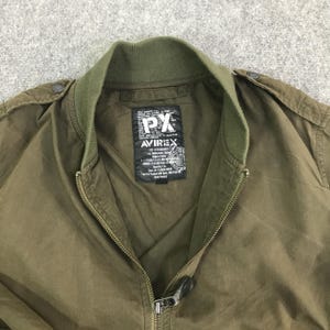 May include: An olive green bomber jacket with a ribbed collar and a full-length front zipper. A black rectangular label is sewn inside the neck area, featuring the white text PX and AVIREX, along with detailed military-style specification text.