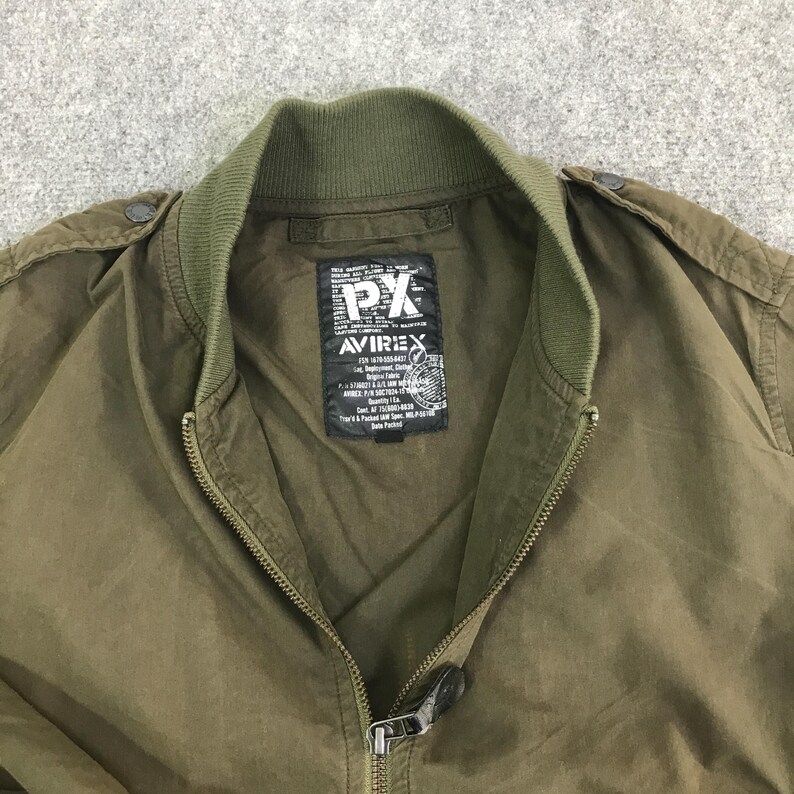 May include: An olive green bomber jacket with a ribbed collar and a full-length front zipper. A black rectangular label is sewn inside the neck area, featuring the white text PX and AVIREX, along with detailed military-style specification text.