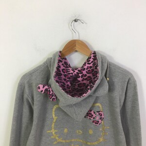 May include: A gray hooded sweatshirt with a gold glitter Hello Kitty design. The hood lining and bow accents are pink with a black leopard print. The sweatshirt is hanging on a wooden hanger.