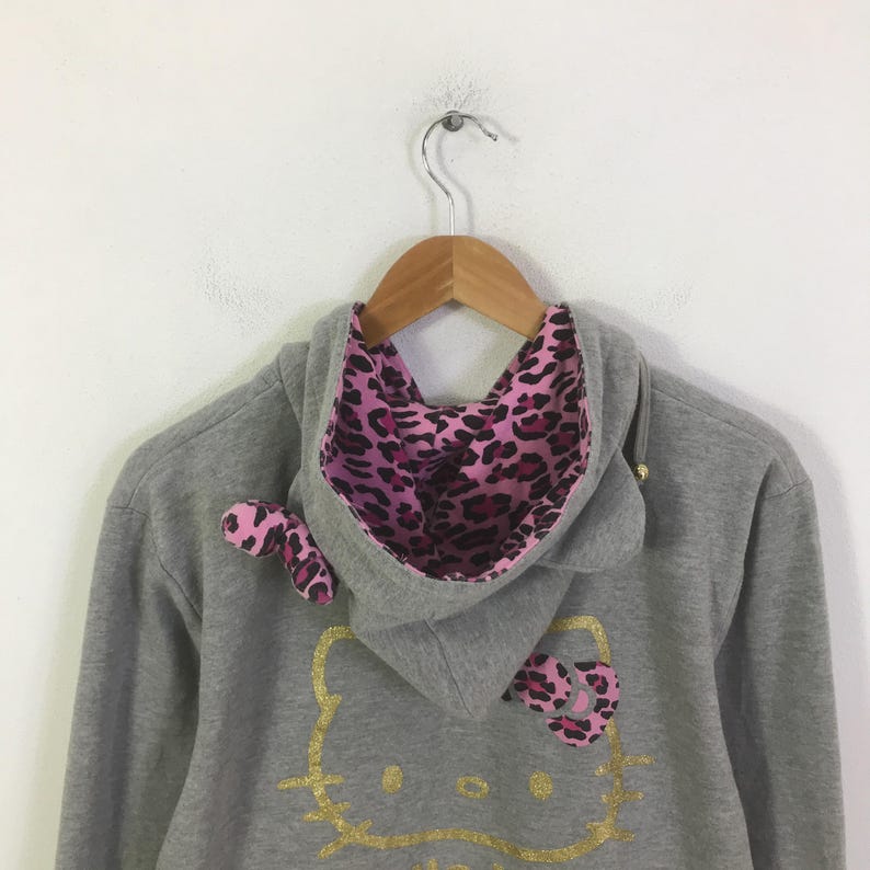 May include: A gray hooded sweatshirt with a gold glitter Hello Kitty design. The hood lining and bow accents are pink with a black leopard print. The sweatshirt is hanging on a wooden hanger.
