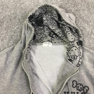 May include: A gray zip-up hoodie with a black and white Hello Kitty pattern lining the hood. The hoodie features a Hello Kitty logo on the front and a skull and crossbones design. The hoodie is made of a soft, comfortable material.