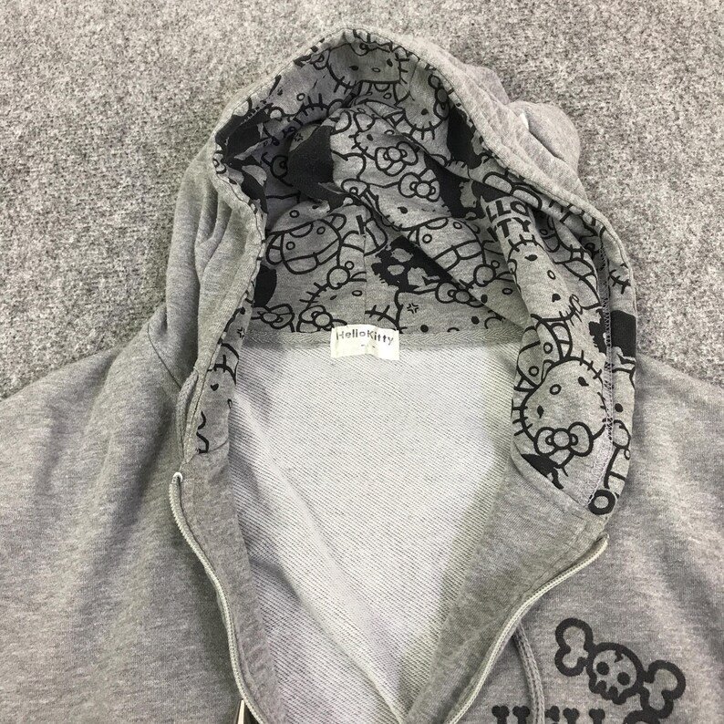 May include: A gray zip-up hoodie with a black and white Hello Kitty pattern lining the hood. The hoodie features a Hello Kitty logo on the front and a skull and crossbones design. The hoodie is made of a soft, comfortable material.