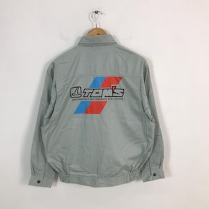 May include: A gray workwear jacket with a red, blue, and white TOM'S logo on the back. The logo reads "TOM'S PERFORMANCE ENGINEERING FOR TOYOTA".
