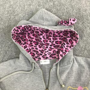 May include: A gray zip-up hoodie with a pink and black leopard print lining in the hood. The hood has a bow on top. The hoodie has a Hello Kitty logo on the front.