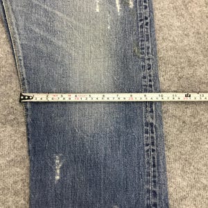 May include: Close-up of light blue denim jeans with a measuring tape. The tape shows measurements in inches, with the jeans' width being approximately 6 inches. The jeans have a distressed look with visible wear and tear.