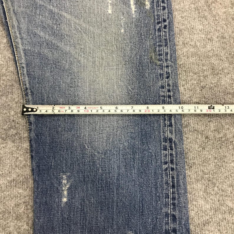 May include: Close-up of light blue denim jeans with a measuring tape. The tape shows measurements in inches, with the jeans' width being approximately 6 inches. The jeans have a distressed look with visible wear and tear.