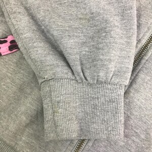 May include: Close-up of a gray sweatshirt sleeve with a ribbed cuff. A pink and brown patterned detail is visible near the zipper. The zipper is gold-colored. The fabric shows signs of wear.