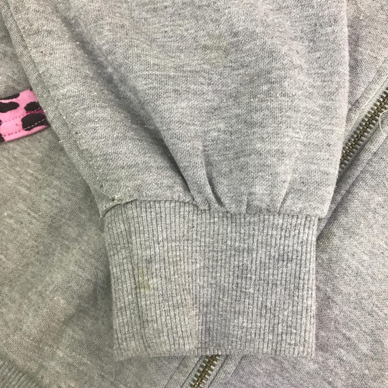 May include: Close-up of a gray sweatshirt sleeve with a ribbed cuff. A pink and brown patterned detail is visible near the zipper. The zipper is gold-colored. The fabric shows signs of wear.